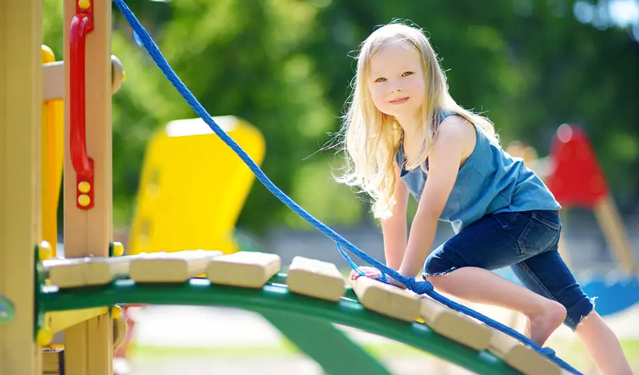 Liten flicka leker på lekplats i solsken, med grönska i bakgrunden, tittar rakt in i kameran